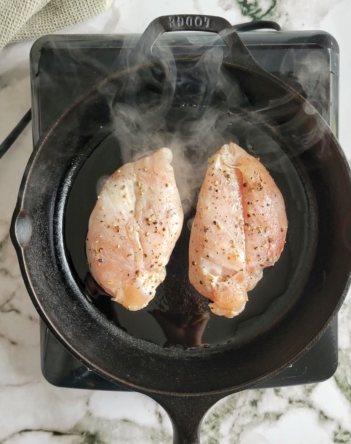 two raw seasoned chicken breasts searing on a cast iron skillet with smoke coming out