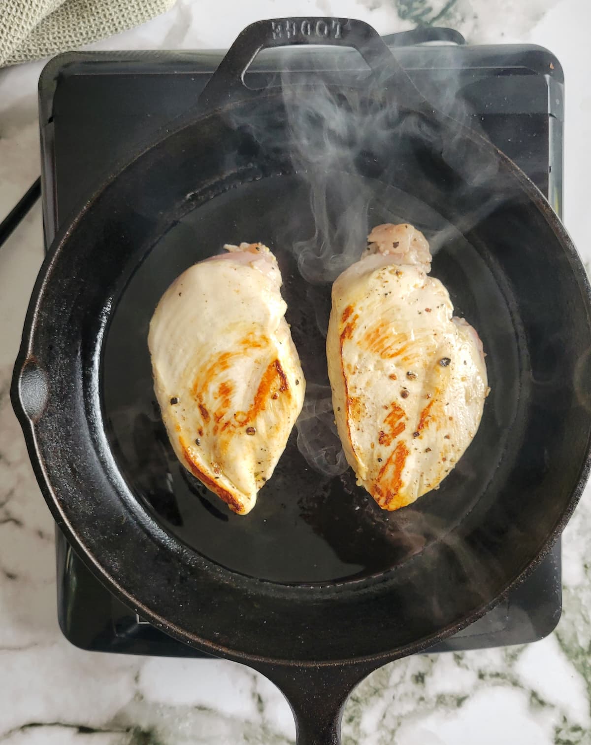 two seasoned chicken breasts searing in a cast iron skillet, smoke coming off