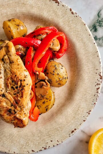 chicken breast on a plate over baby potatoes, mushrooms and sliced red bell peppers. lemon on the side