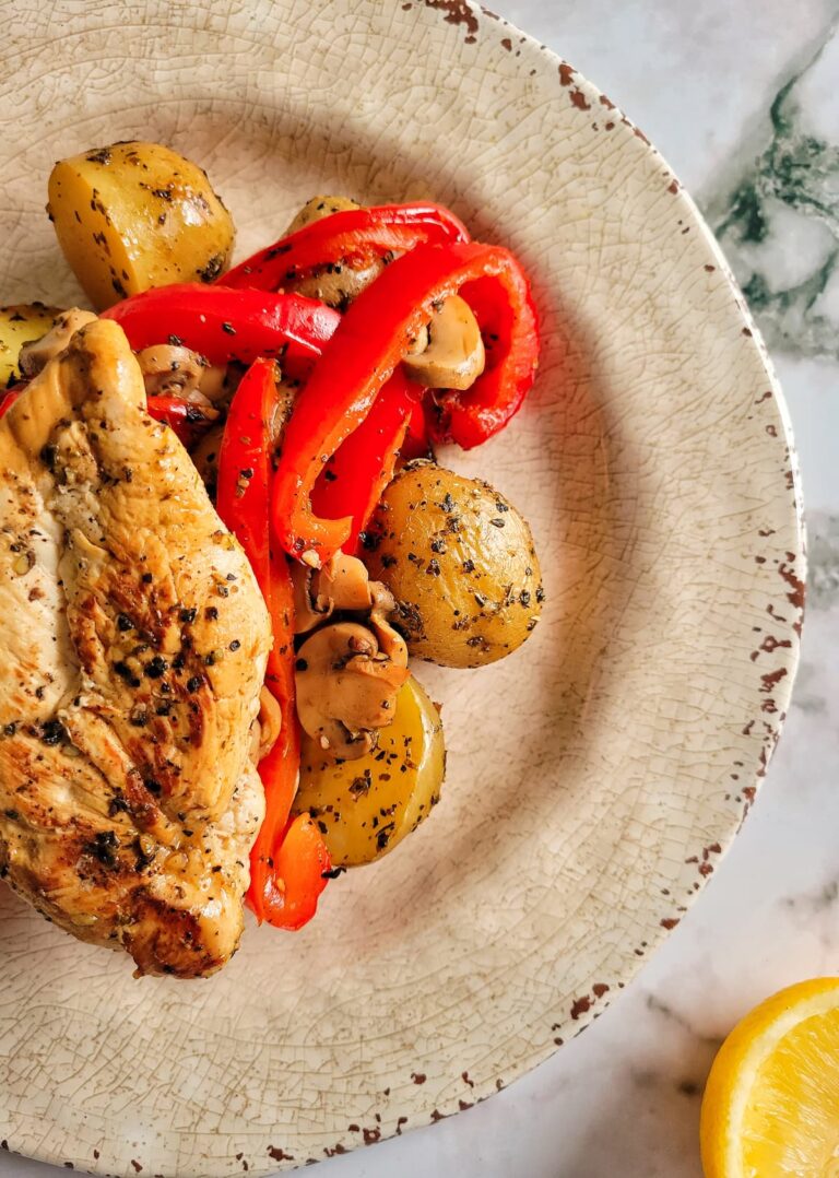 chicken breast on a plate over baby potatoes, mushrooms and sliced red bell peppers. lemon on the side