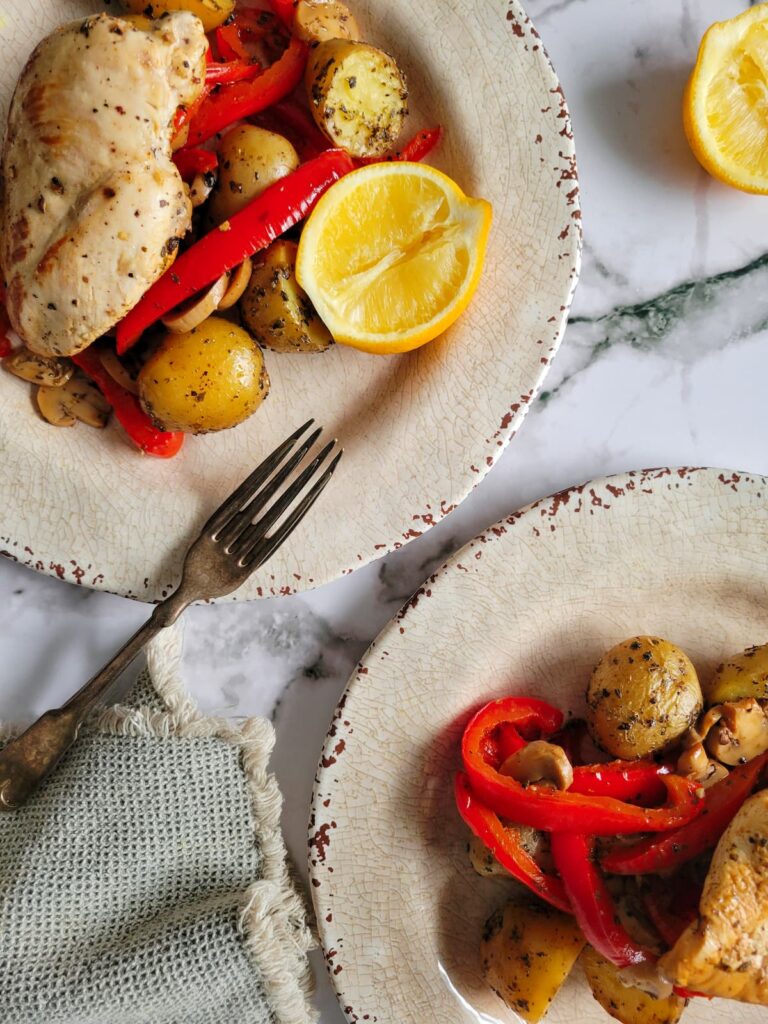 two plates of chicken, potatoes, red bell peppers and halved lemons, fork on one plate