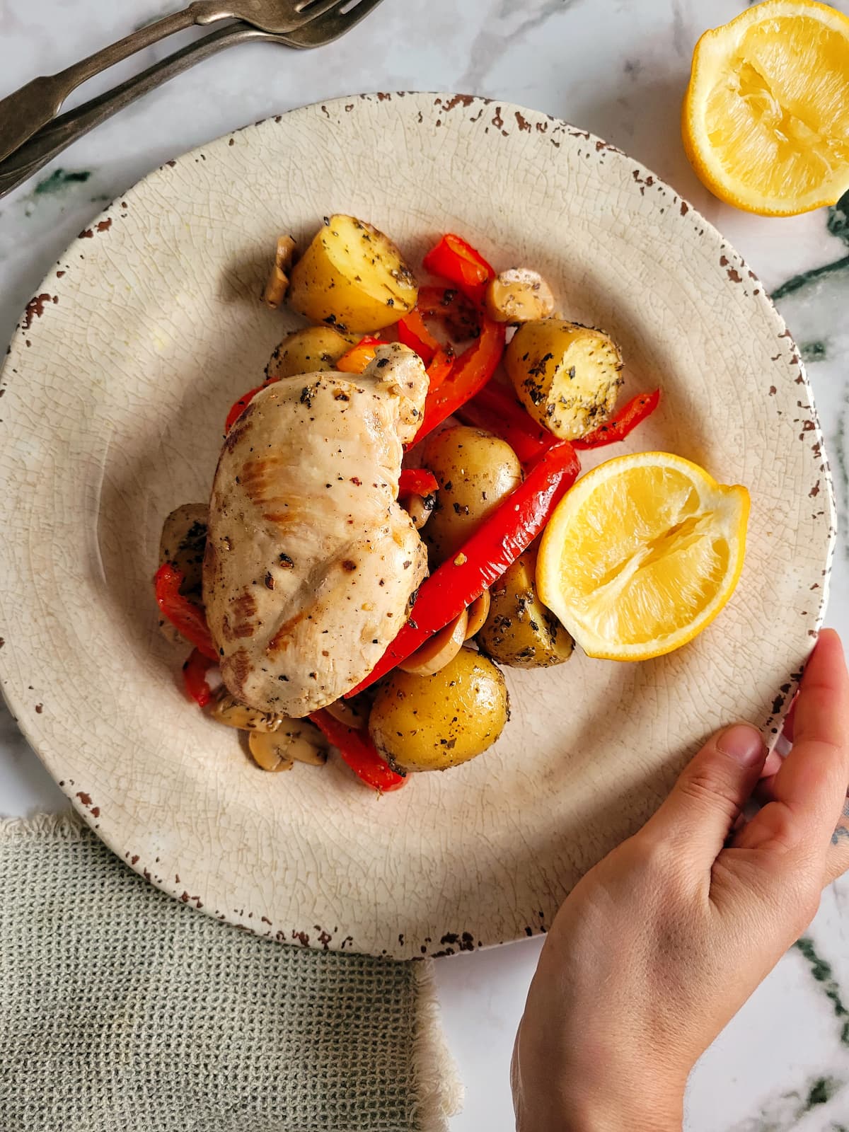hand holding a plate with a chicken breast over potatoes, mushroom and red bell pepper, halved lemons on and around the plate