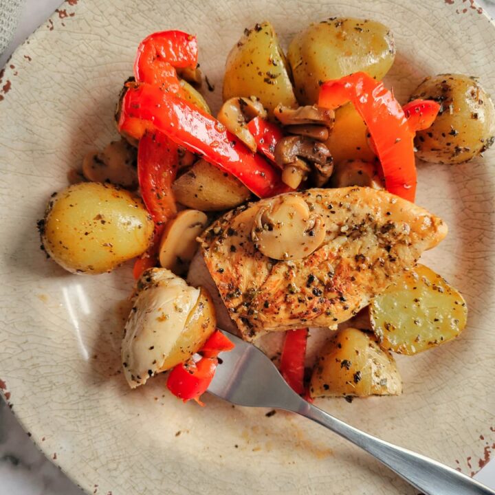 sliced seasoned chicken on a plate with baby potatoes, mushrooms and red bell peppers, fork piercing some of the chicken