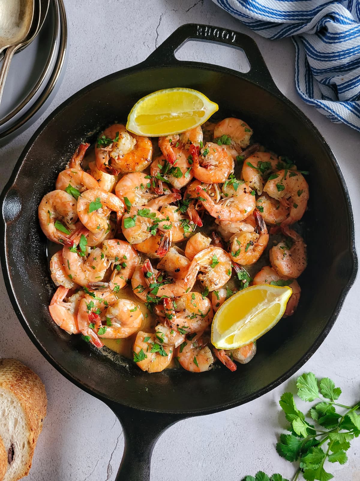 cast iron skillet with sauteed shrimp garnished with fresh chopped parsley and lemon wedges, bread and plates in the background