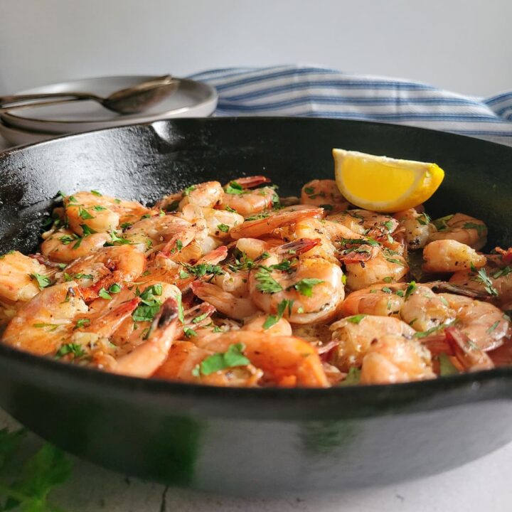 side view of sauteed shrimp in a cast iron skillet garnished with fresh chopped parsley and a lemon wedge