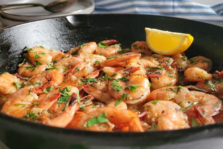 side view of sauteed shrimp in a cast iron skillet garnished with fresh chopped parsley and a lemon wedge