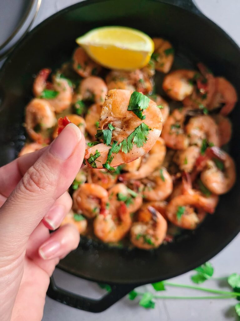 hand holding a sauteed shrimp with fresh parsley over a skillet with the rest, lemon wedge in the skillet