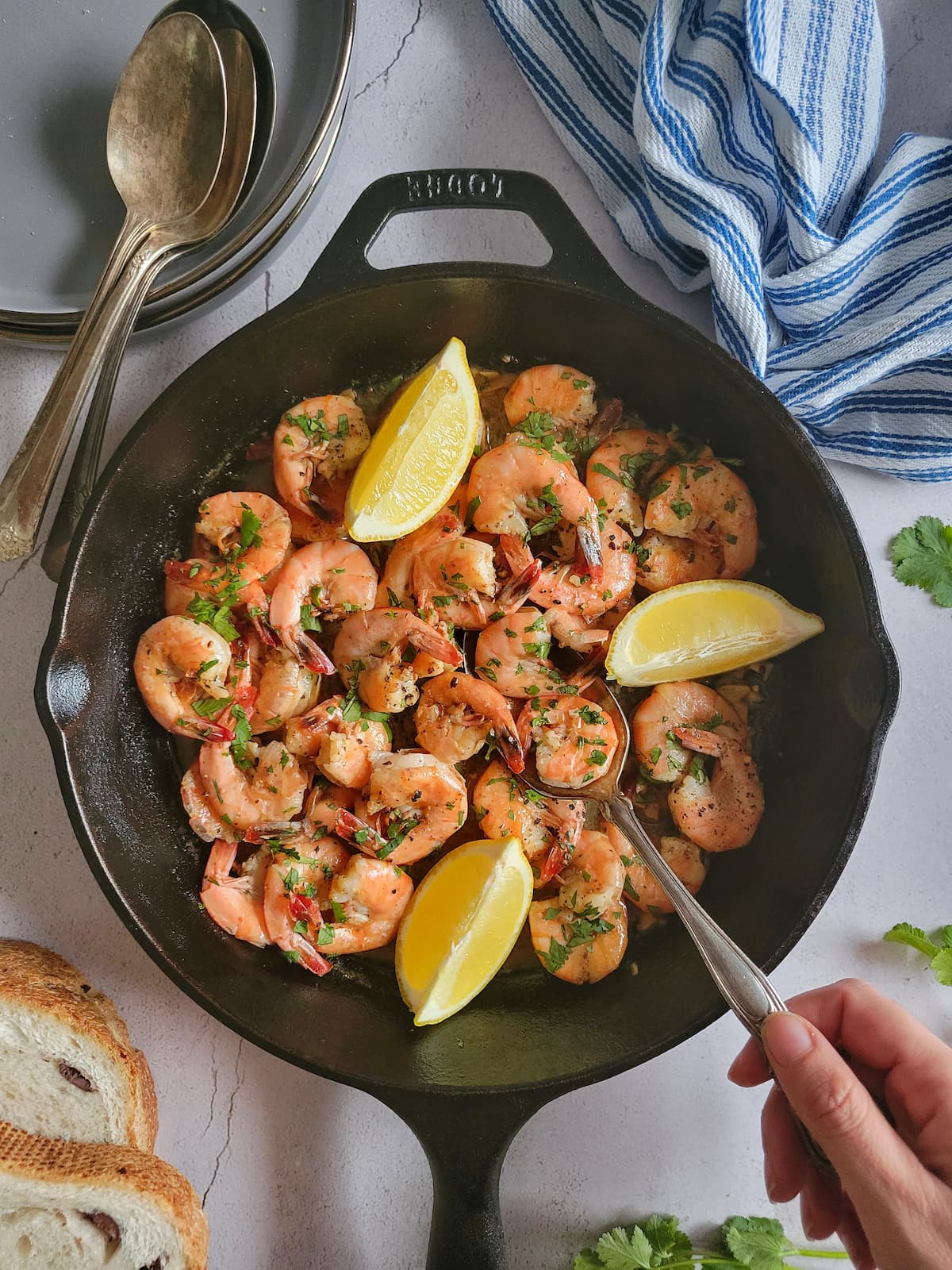 cooked shell on shrimp in a cast iron pan garnished with fresh chopped parsley and 3 lemon wedges, spoon with a hand in the skillet, bread, plates and more spoons in the background
