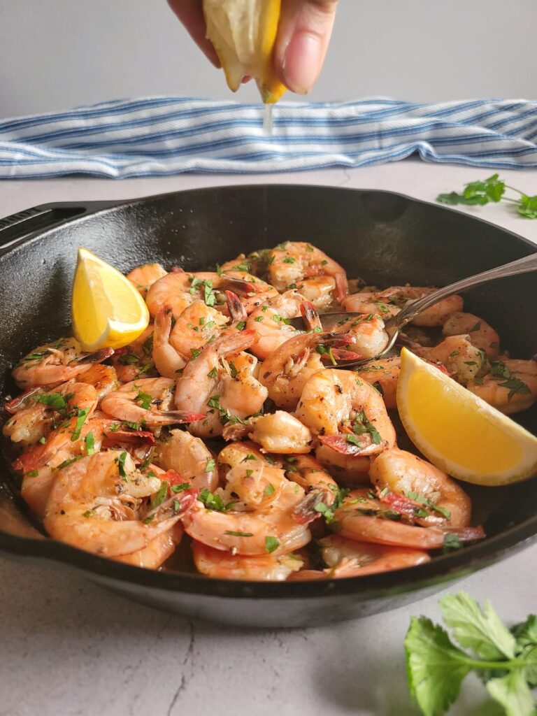 hand squeezing a lemon wedge over a skillet of cooked shell on shrimp garnished with fresh parsley, more lemon and a spoon in the skillet