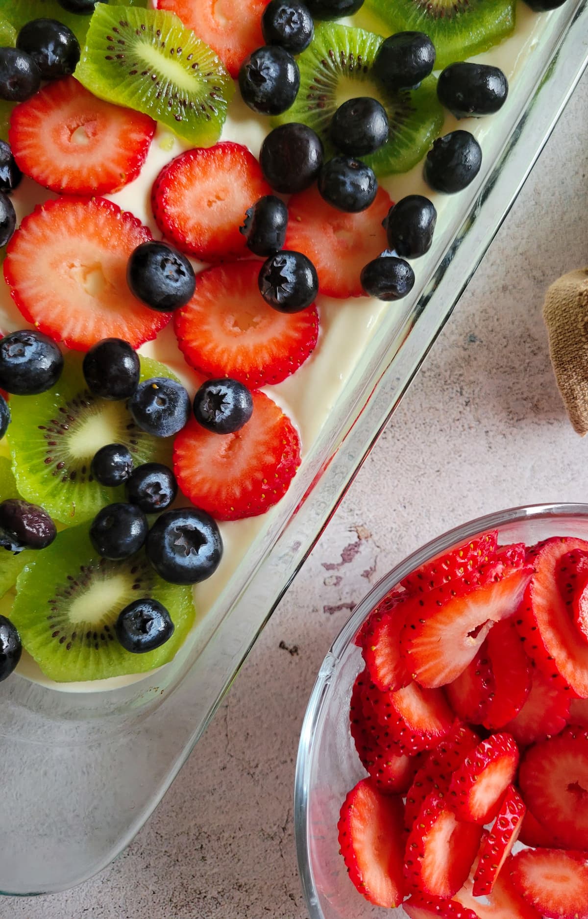 bowl of sliced fresh strawberries next to a glass baking dish with a dessert topped with fresh blueberries, sliced kiwis and strawberries