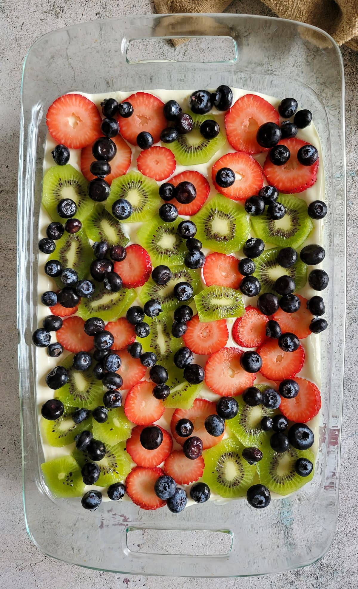 sliced kiwi, strawberries and blueberries on top of cream in a glass baking dish
