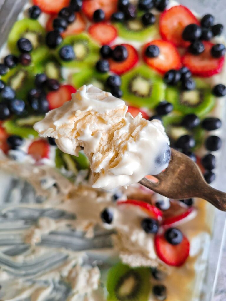 fork with layered crackers and whipped cream over a baking dish with a dessert topped with fresh blueberries, strawberries and kiwis