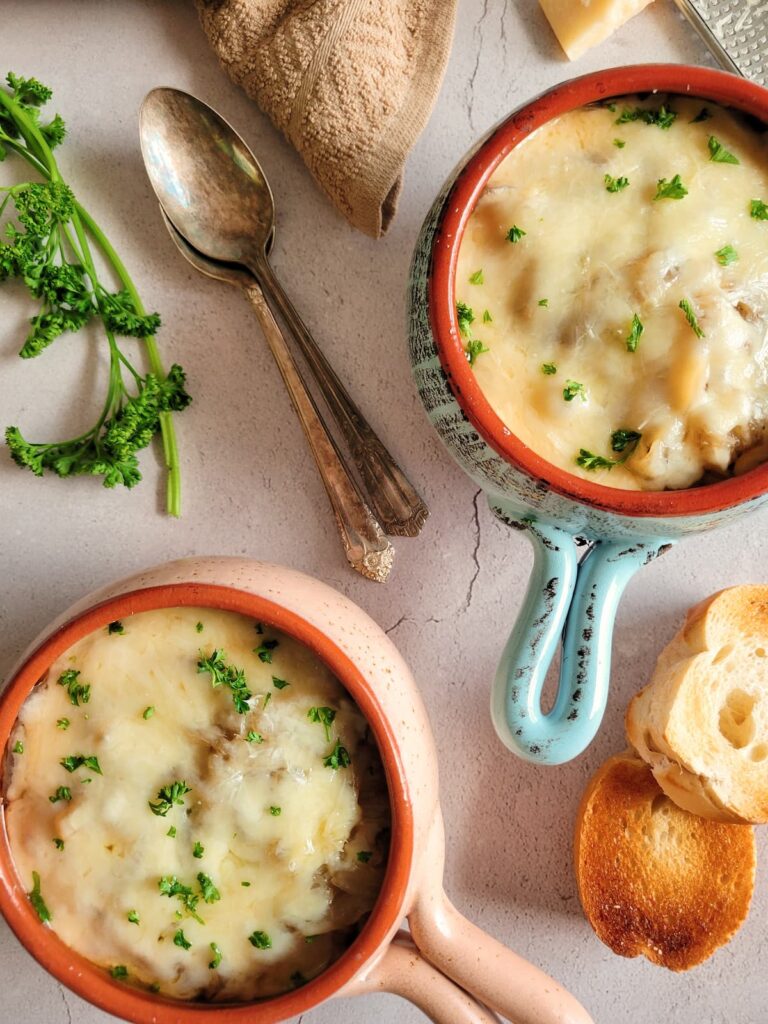 two bowls of french onion soup topped with fresh parsley, sliced bread, spoons and fresh herbs in the background