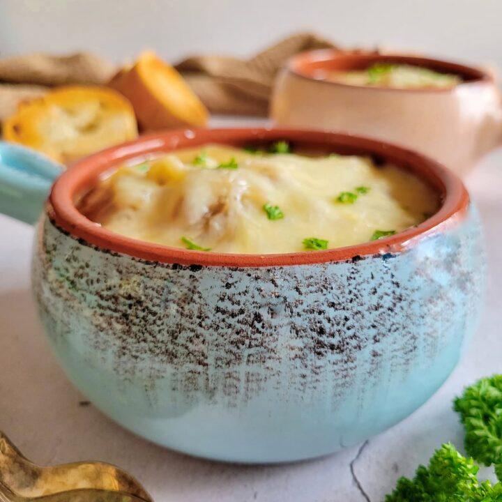 side view of a bowl of french onion soup topped with fresh chopped parsley and melted cheese, more herbs, another bowl of soup, sliced bread and spoons in the background