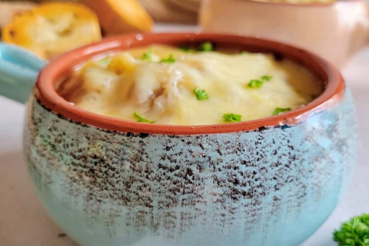 side view of a bowl of french onion soup topped with fresh chopped parsley and melted cheese, more herbs, another bowl of soup, sliced bread and spoons in the background