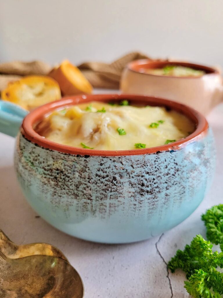 side view of a bowl of french onion soup topped with fresh chopped parsley and melted cheese, more herbs, another bowl of soup, sliced bread and spoons in the background