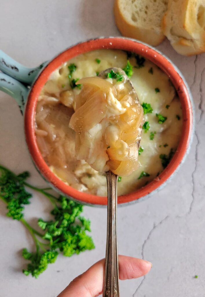 hand holding up a spoonful of french onion soup over a bowl with the rest of it, fresh herbs and sliced bread in the background