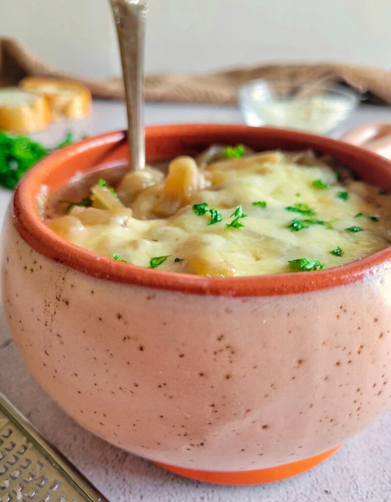 spoon in a bowl of french onion soup topped with melted cheese and fresh chopped parsley, sliced bread in the background