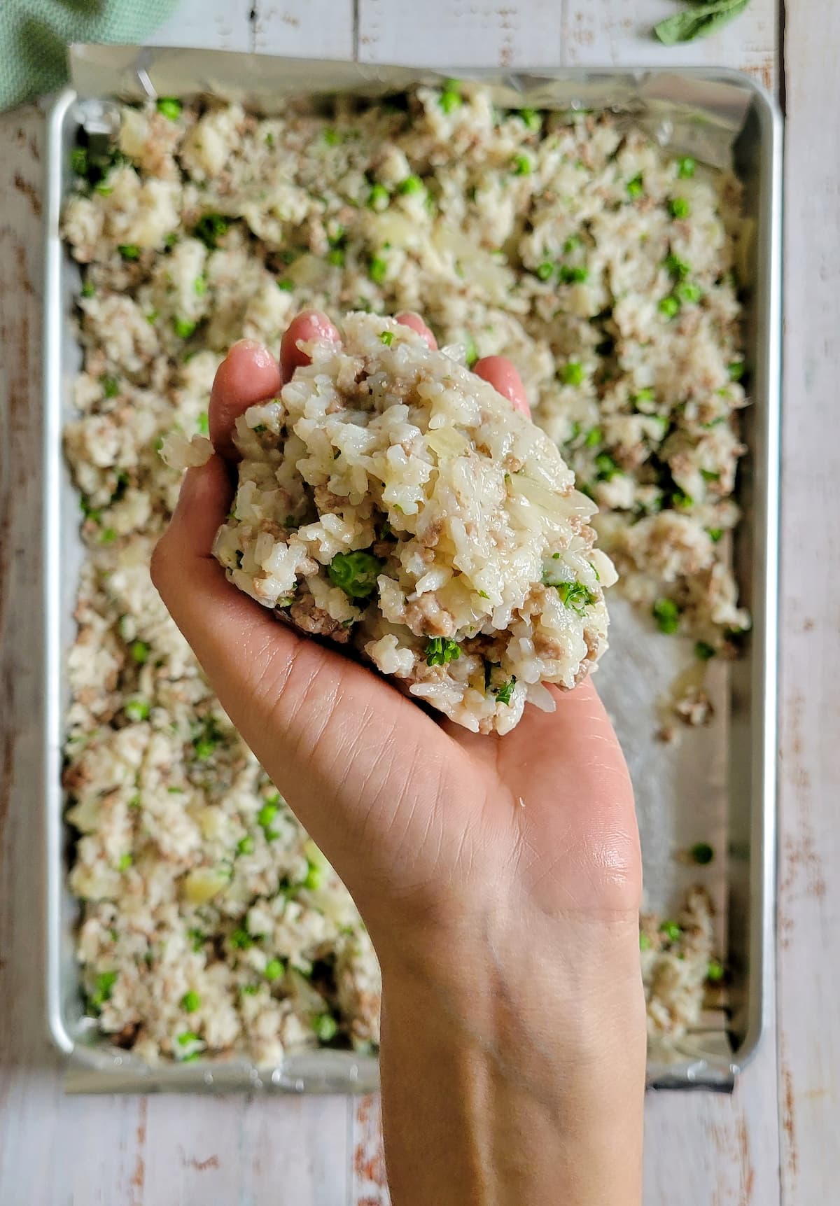 hand holding a ball of rice, ground beef and peas over a sheet pan of the rest of the rice mixture spread out