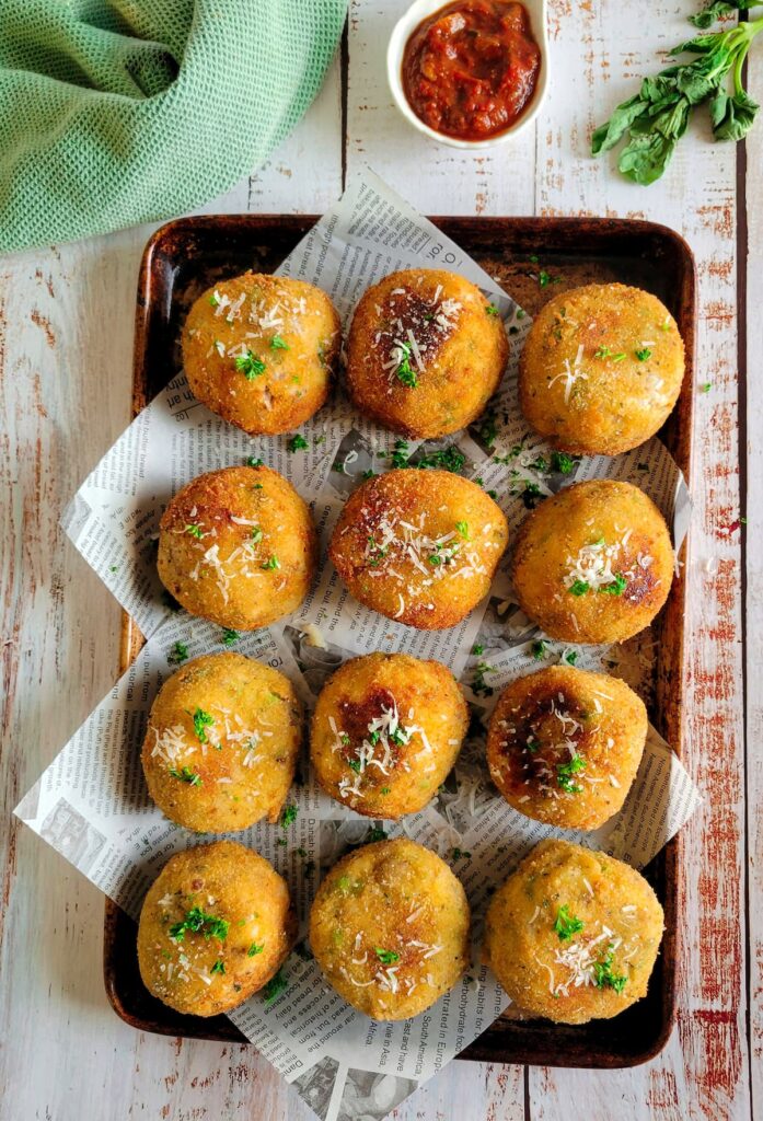 12 crispy arancini balls on a lined baking sheet topped with fresh parsley and grated parmesan cheese, ramekin of tomato sauce and fresh basil in the background