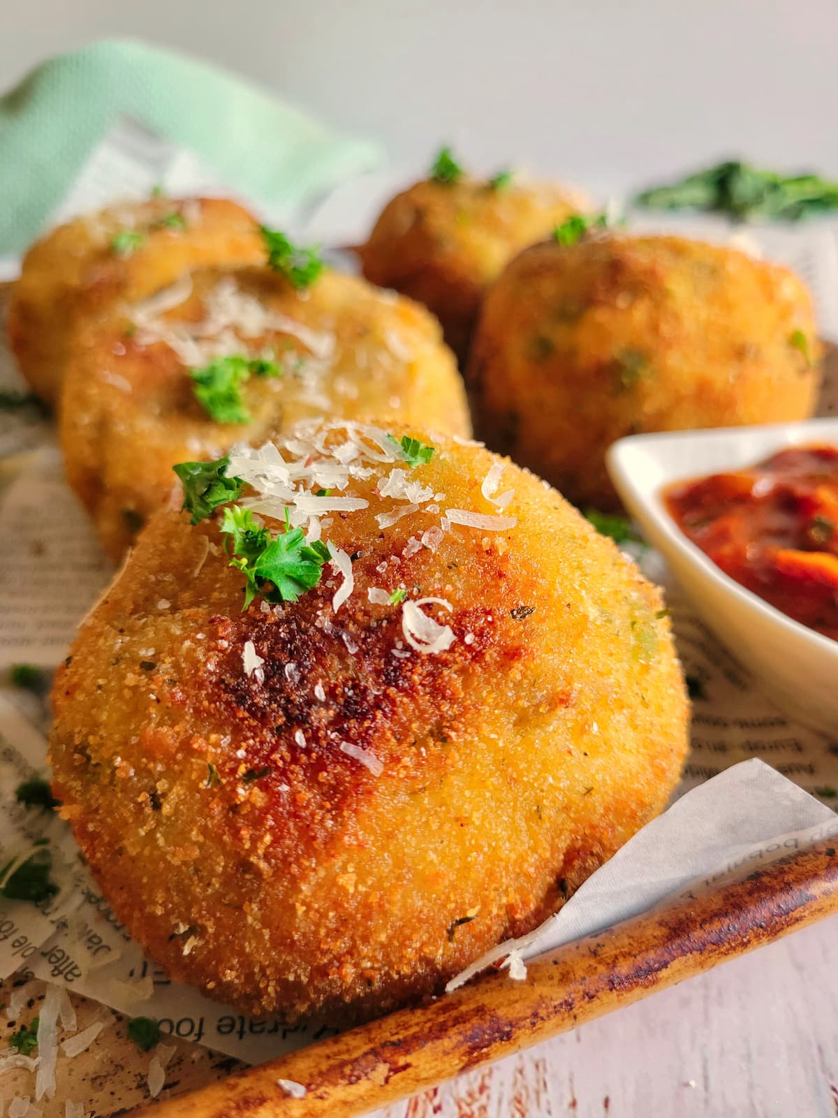 close up of a crispy rice ball garnished with fresh parsley and parmesan cheese on a sheet pan with the rest of them, tomato sauce on the tray
