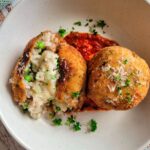 two arancini with tomato sauce on a plate garnished with fresh parsley and grated parmesan cheese, one of the rice balls is split open to show the rice, cheese and peas in the center