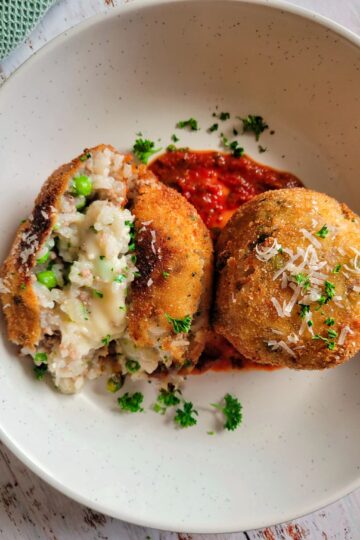 two arancini with tomato sauce on a plate garnished with fresh parsley and grated parmesan cheese, one of the rice balls is split open to show the rice, cheese and peas in the center