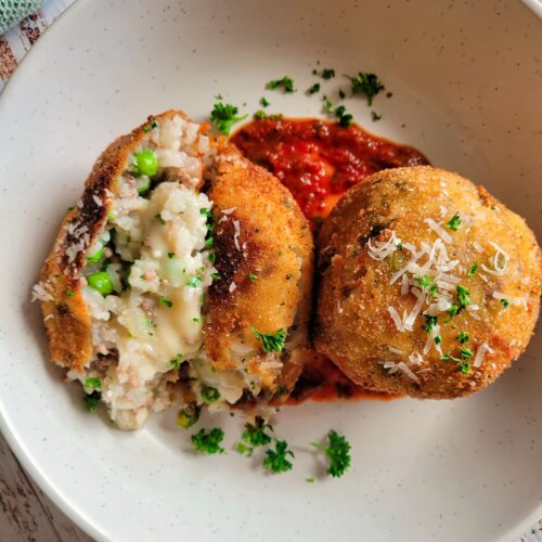 two arancini with tomato sauce on a plate garnished with fresh parsley and grated parmesan cheese, one of the rice balls is split open to show the rice, cheese and peas in the center