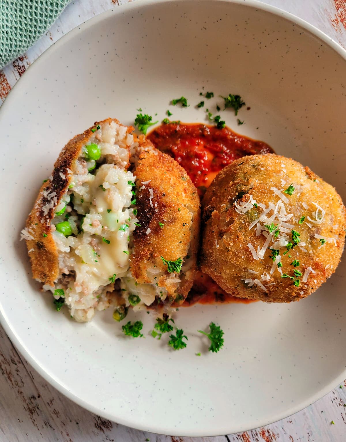 two arancini with tomato sauce on a plate garnished with fresh parsley and grated parmesan cheese, one of the rice balls is split open to show the rice, cheese and peas in the center