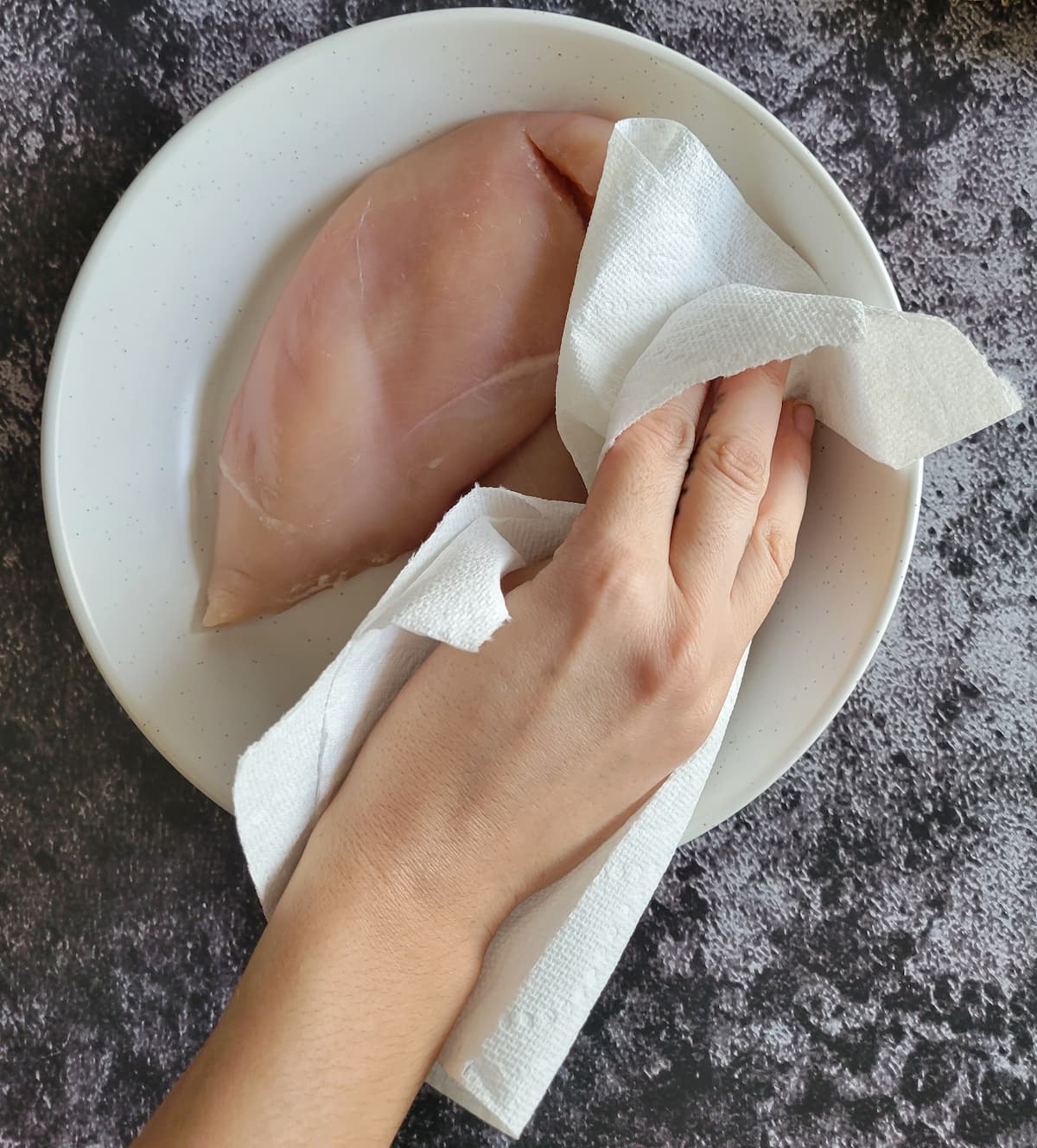 hand with a paper towel patting down raw chicken breasts in a bowl