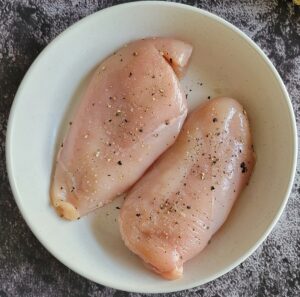 two raw chicken breasts in a bowl seasoned with salt and pepper