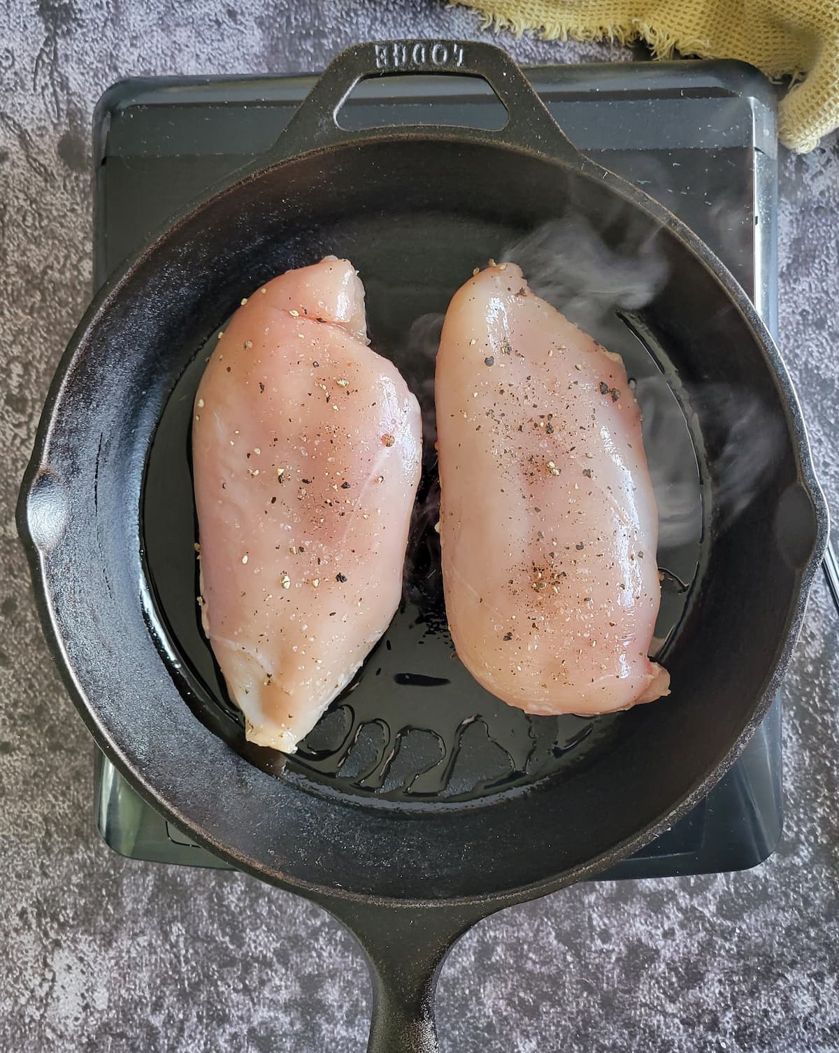 two raw chicken breasts seasoned with salt and pepper searing in a cast iron skillet on a burner