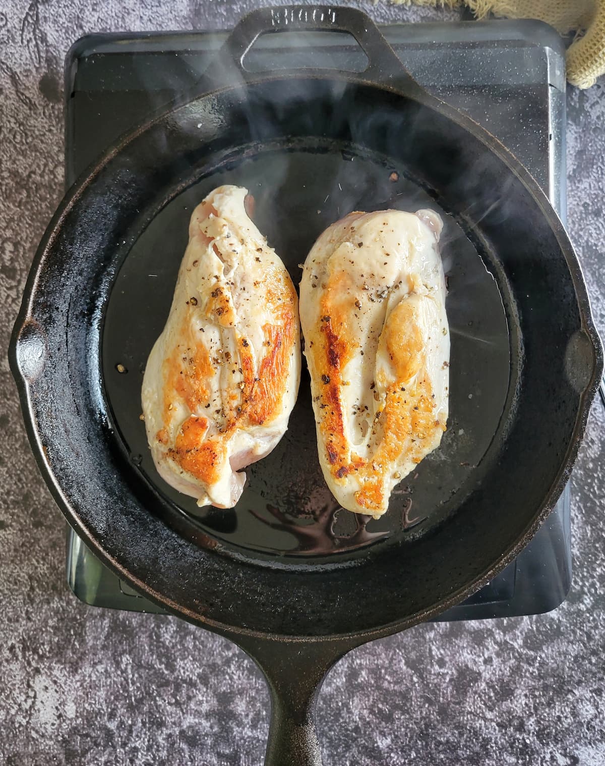 two seasoned chicken breasts searing in a cast iron skillet on a burner, tops are golden brown