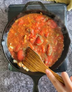 hand with a wooden spatula crushing whole tomatoes in a cast iron skillet with seasonings and diced onions