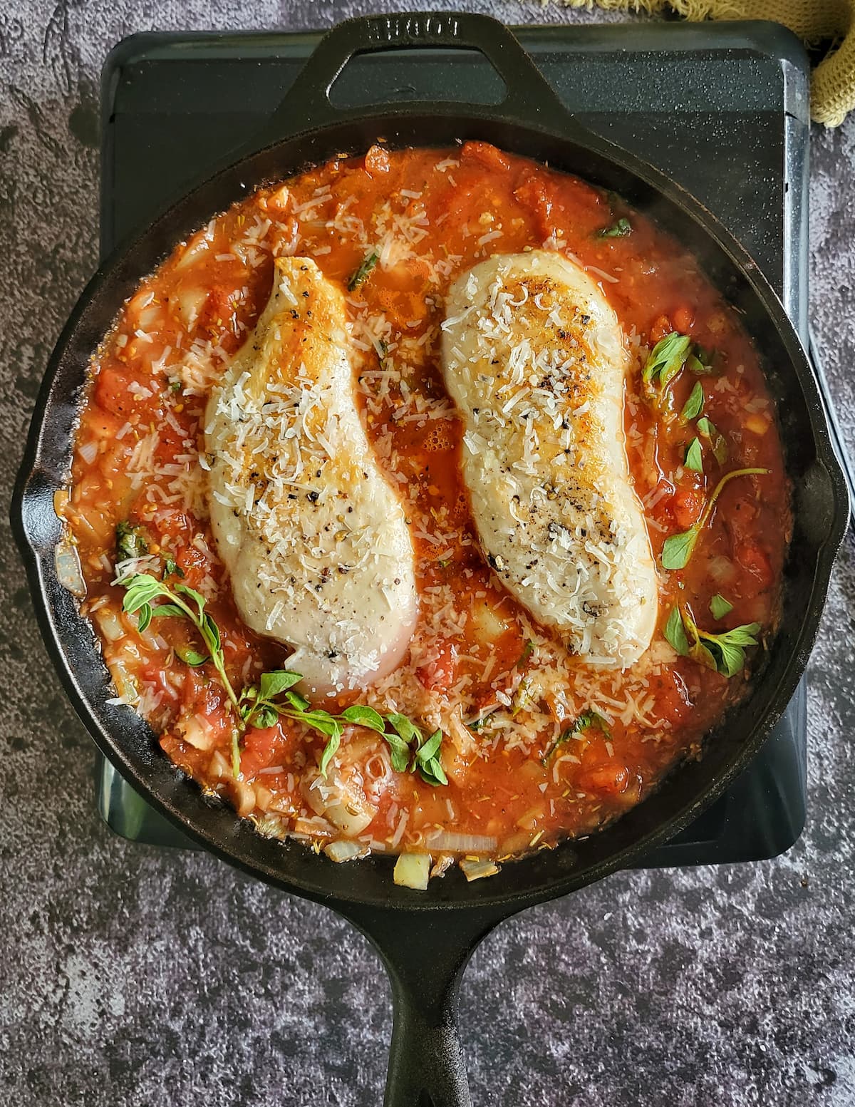 two chicken breasts seasoned with salt, pepper and grated parmesan cheese in a cast iron skillet with tomato sauce and sprigs of fresh oregano