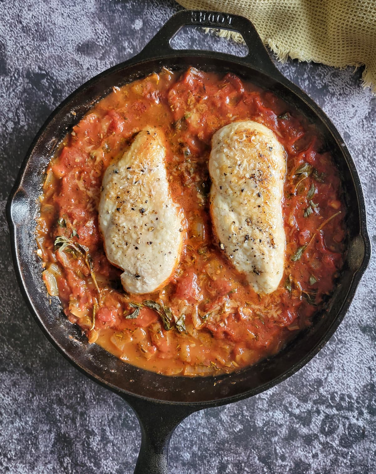 two cooked and seasoned chicken breasts in tomato sauce with herbs in a cast iron skillet