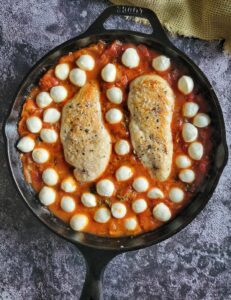 two cooked and seasoned chicken breasts in tomato sauce surrounded by mini bocconcini balls in a cast iron skillet