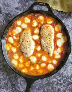 two cooked and seasoned chicken breasts in tomato sauce surrounded by melted mini bocconcini balls in a cast iron skillet
