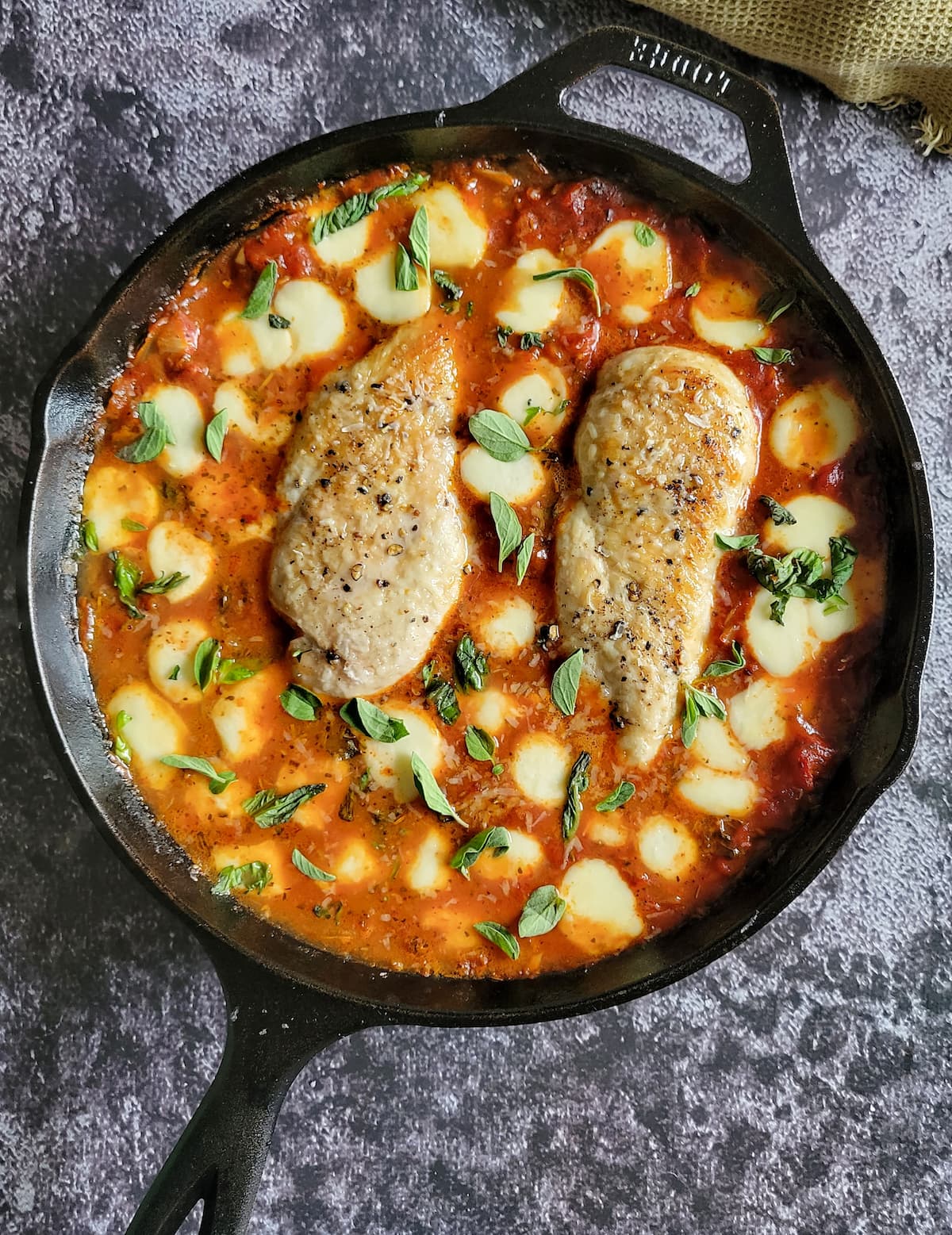 two cooked and seasoned chicken breasts in tomato sauce surrounded by melted mini bocconcini balls and garnished with fresh chopped basil in a cast iron skillet