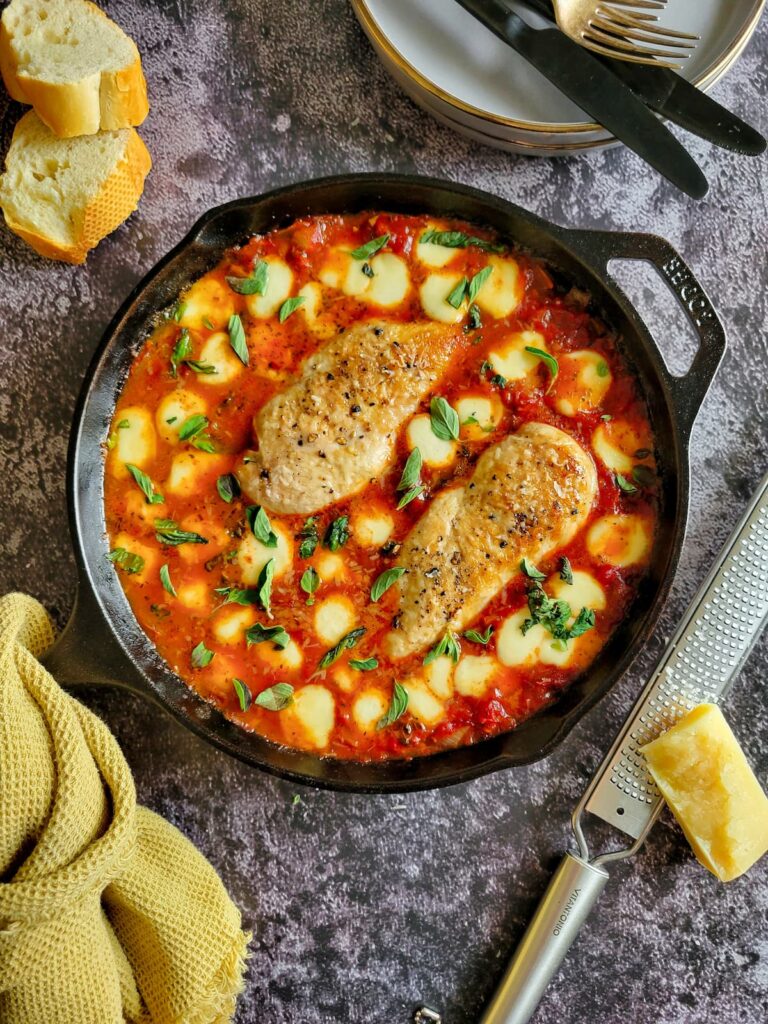 two cooked and seasoned chicken breasts in tomato sauce surrounded by melted bocconcini and garnished with chopped fresh basil in a cast iron skillet, sliced bread in the background with plates with forks and knifes and cheese next to a microplane
