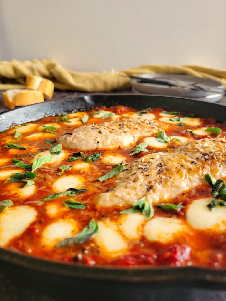 side view of two cooked and seasoned chicken breasts in tomato sauce surrounded by melted bocconcini and garnished with chopped fresh basil in a cast iron skillet, bread in the background