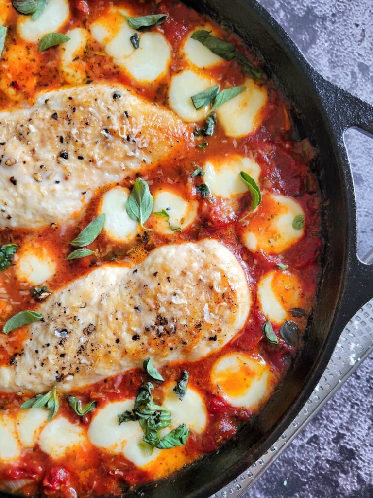 close up of two cooked and seasoned chicken breasts in tomato sauce surrounded by melted bocconcini and garnished with chopped fresh basil in a cast iron skillet