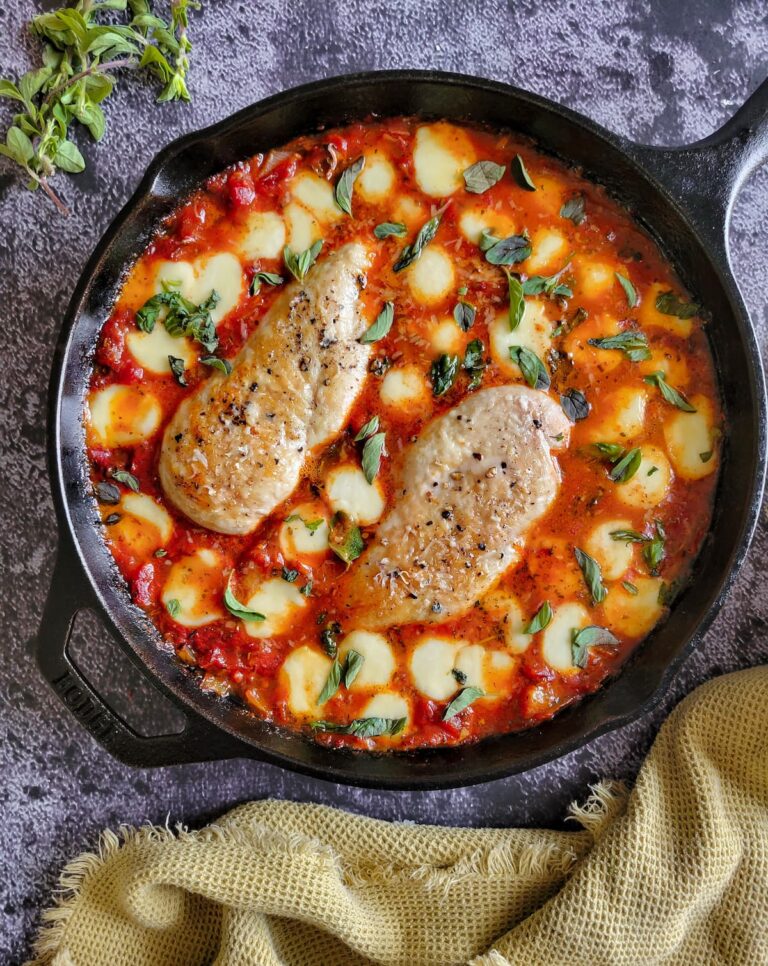 two cooked and seasoned chicken breasts in tomato sauce surrounded by melted bocconcini and garnished with chopped fresh basil in a cast iron skillet, fresh herbs in the background