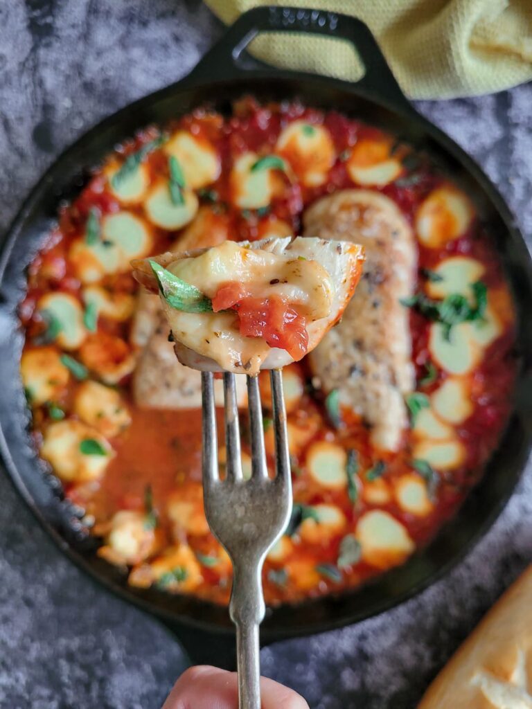 fork piercing a piece of chicken breast in tomato sauce with melted bocconcini and chopped fresh basil over a cast iron skillet with the rest of it