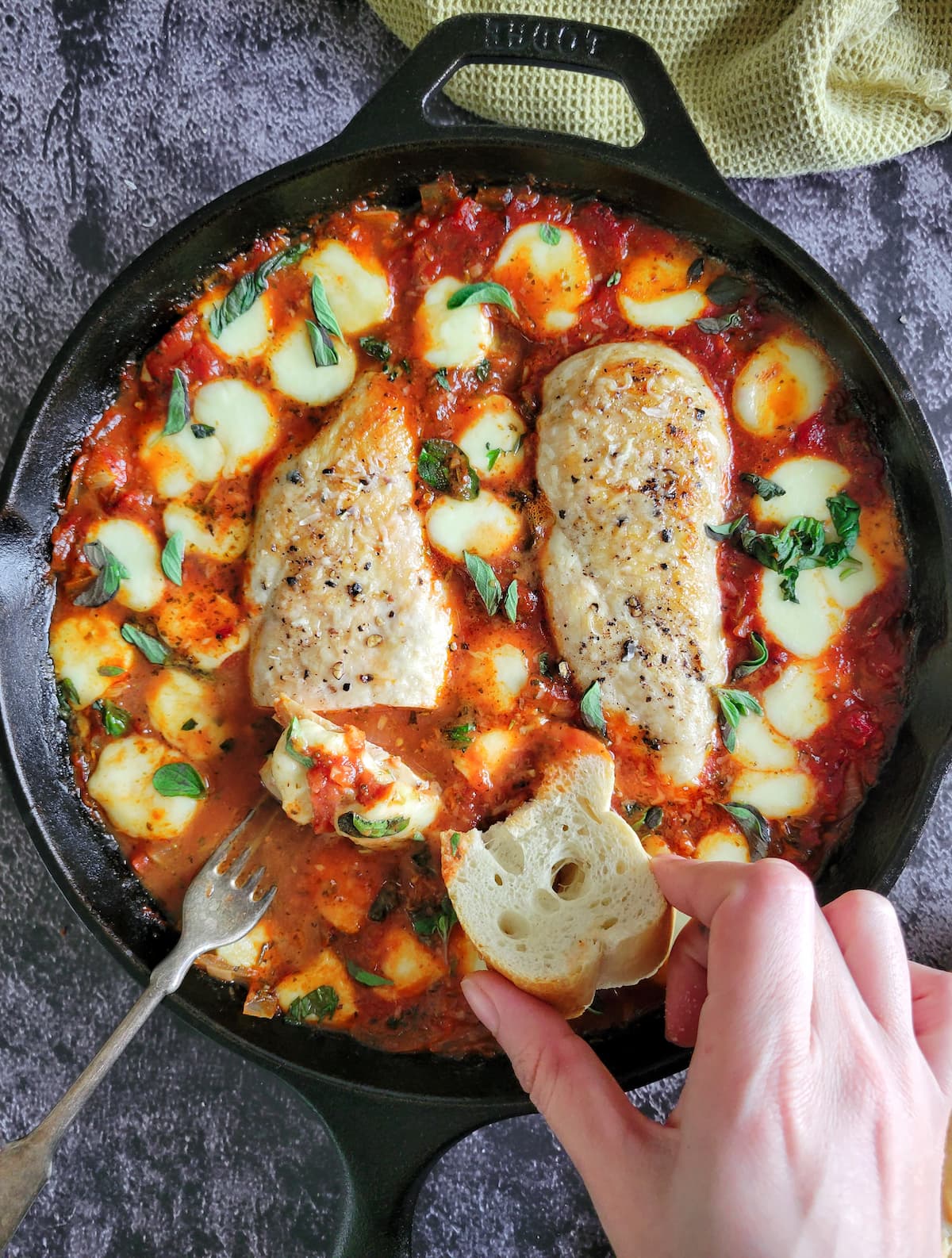 hand with a slice of bread in a cast iron skillet with tomato sauce, melted bocconcini, chopped fresh basil and two seasoned cooked chicken breasts