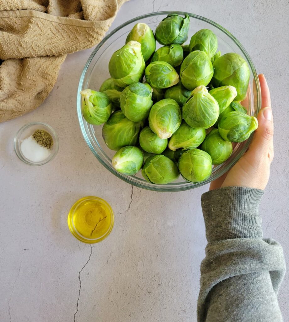 ramekin of salt and pepper, ramekin of olive oil next to a bowl of brussels sprouts, hand on the bowl