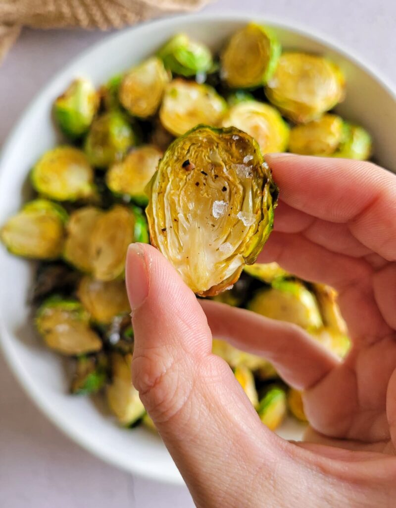 hand holding a crispy brussels sprout garnished with flakey sea salt over a bowl of the rest of them