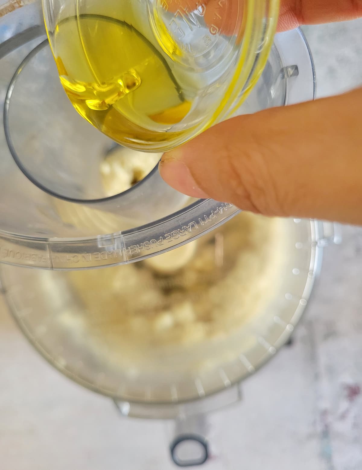 hand pouring a ramekin of oil into the mouth of a food processor