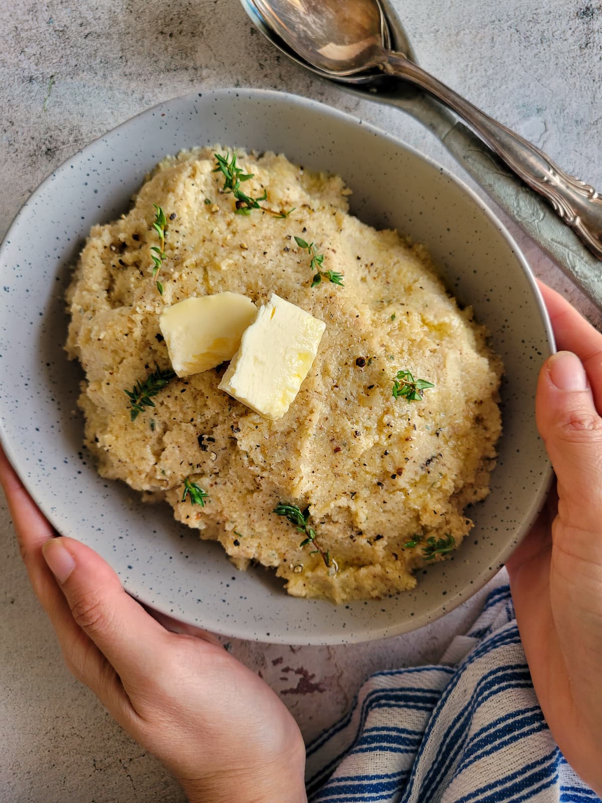 hands holding a bowl of mashed cauliflower with 2 pats of butter and fresh thyme, two spoons on the side of the bowl