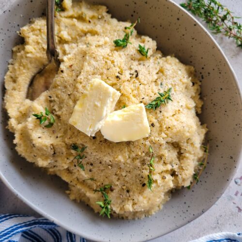 cauliflower mashed potatoes in a bowl topped with butter and fresh thyme, more thyme in the background, spoon in the bowl
